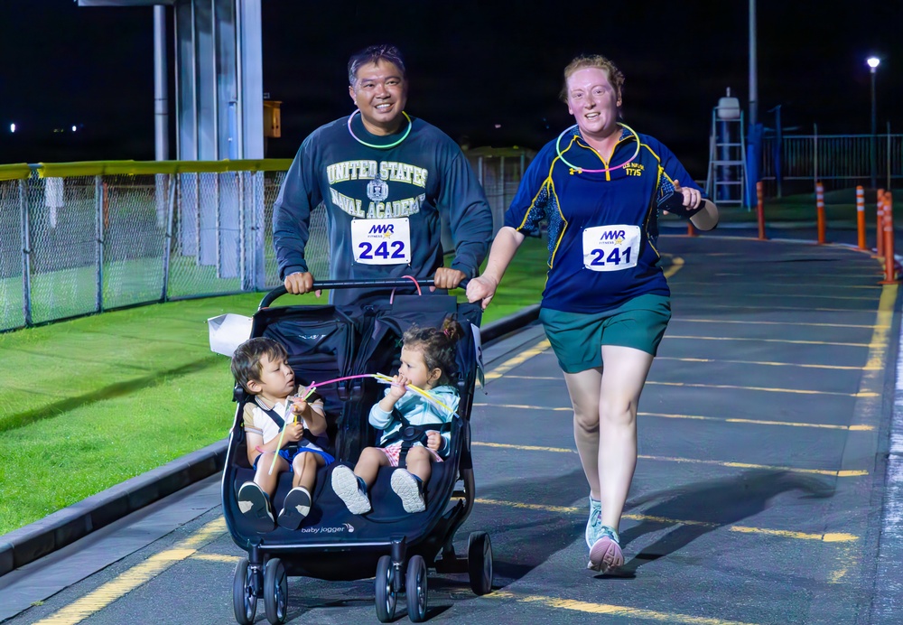 A family participates in a nighttime MWR (Morale, Welfare, and Recreation) running race on what appears to be a military installation. A man wearing a United States Naval Academy sweatshirt pushes a Baby Jogger double stroller containing two young children, while a woman running alongside him wears race bibs numbered 241 and 242. The scene is well-lit by artificial lighting against a dark night sky.