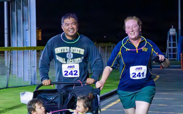 A family participates in a nighttime MWR (Morale, Welfare, and Recreation) running race on what appears to be a military installation. A man wearing a United States Naval Academy sweatshirt pushes a Baby Jogger double stroller containing two young children, while a woman running alongside him wears race bibs numbered 241 and 242. The scene is well-lit by artificial lighting against a dark night sky.