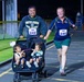 A family participates in a nighttime MWR (Morale, Welfare, and Recreation) running race on what appears to be a military installation. A man wearing a United States Naval Academy sweatshirt pushes a Baby Jogger double stroller containing two young children, while a woman running alongside him wears race bibs numbered 241 and 242. The scene is well-lit by artificial lighting against a dark night sky.