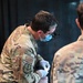 A U.S. military medical personnel in OCP (Operational Camouflage Pattern) uniform is performing a medical procedure on a patient. The provider is wearing magnification loupes, a surgical mask, and latex gloves, suggesting a clinical or field medical setting. A second uniformed individual is observing in the background.
