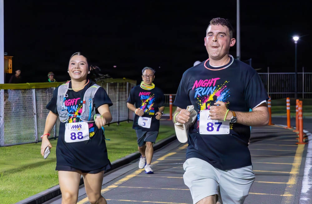 Three participants are running in a nighttime race event called 'Night Run,' organized by MWR (Morale, Welfare and Recreation). The runners are wearing matching dark navy 'Night Run' t-shirts with colorful graphics and numbered race bibs. The event takes place on a paved track or road under artificial lighting.
