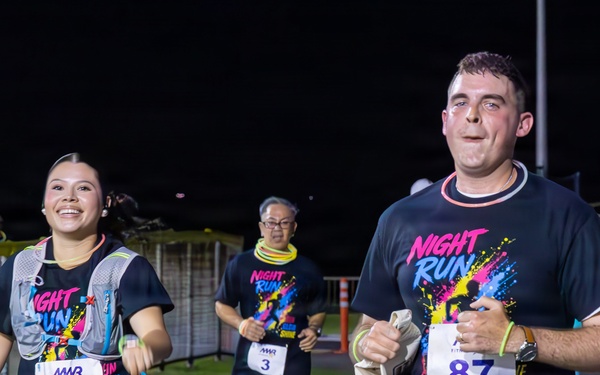 Three participants are running in a nighttime race event called 'Night Run,' organized by MWR (Morale, Welfare and Recreation). The runners are wearing matching dark navy 'Night Run' t-shirts with colorful graphics and numbered race bibs. The event takes place on a paved track or road under artificial lighting.