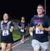 Three participants are running in a nighttime race event called 'Night Run,' organized by MWR (Morale, Welfare and Recreation). The runners are wearing matching dark navy 'Night Run' t-shirts with colorful graphics and numbered race bibs. The event takes place on a paved track or road under artificial lighting.