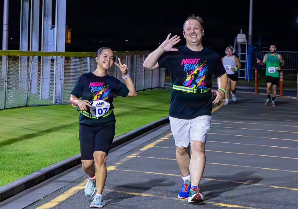 Two runners in matching black 'Night Run' t-shirts are participating in a nighttime running event, smiling and gesturing toward the camera. The woman on the left wears race bib number 107 issued by MWR (Morale, Welfare and Recreation), while the man on the right waves with an open hand. Additional runners are visible in the background on what appears to be a paved track or road course.