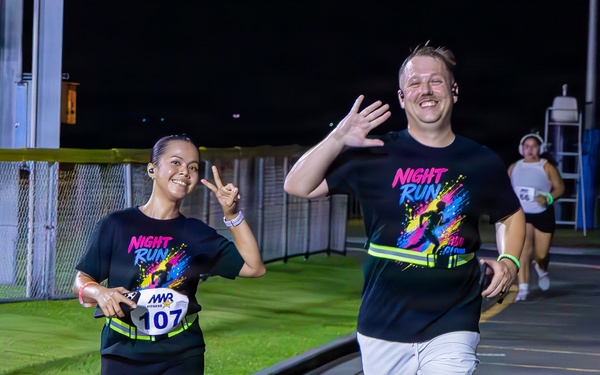 Two runners in matching black 'Night Run' t-shirts are participating in a nighttime running event, smiling and gesturing toward the camera. The woman on the left wears race bib number 107 issued by MWR (Morale, Welfare and Recreation), while the man on the right waves with an open hand. Additional runners are visible in the background on what appears to be a paved track or road course.