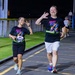 Two runners in matching black 'Night Run' t-shirts are participating in a nighttime running event, smiling and gesturing toward the camera. The woman on the left wears race bib number 107 issued by MWR (Morale, Welfare and Recreation), while the man on the right waves with an open hand. Additional runners are visible in the background on what appears to be a paved track or road course.