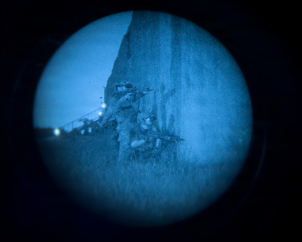 The image appears to be captured through a circular porthole or lens, showing what appears to be an underwater shipwreck scene illuminated in blue tones. The circular vignette framing suggests this may have been photographed through a submarine porthole or underwater vehicle viewport. Debris, structural wreckage, and artificial lighting points are visible against a dark seafloor environment.