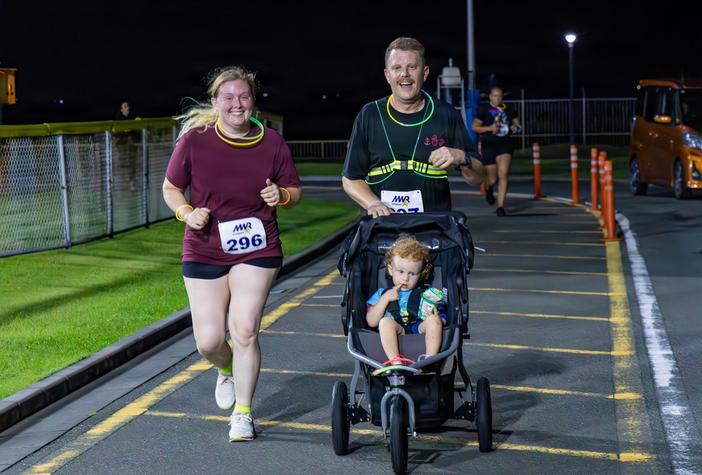 A nighttime road race organized by MWR (Morale, Welfare and Recreation) featuring two adult runners wearing race bibs, one of whom is pushing a jogging stroller with a young toddler seated inside. The runners appear cheerful and energetic, wearing glow accessories typical of night run events. The race takes place on a paved road or parking area with orange traffic cones and fencing visible in the background.