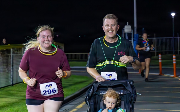 A nighttime road race organized by MWR (Morale, Welfare and Recreation) featuring two adult runners wearing race bibs, one of whom is pushing a jogging stroller with a young toddler seated inside. The runners appear cheerful and energetic, wearing glow accessories typical of night run events. The race takes place on a paved road or parking area with orange traffic cones and fencing visible in the background.