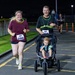 A nighttime road race organized by MWR (Morale, Welfare and Recreation) featuring two adult runners wearing race bibs, one of whom is pushing a jogging stroller with a young toddler seated inside. The runners appear cheerful and energetic, wearing glow accessories typical of night run events. The race takes place on a paved road or parking area with orange traffic cones and fencing visible in the background.