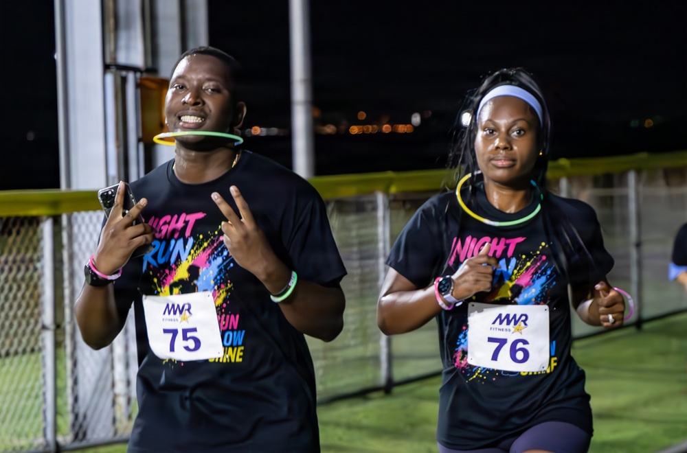 Two runners participating in a nighttime fun run event organized by MWR Fitness, wearing matching 'Night Run' themed black t-shirts with colorful paint splatter designs. Runner on the left wears bib number 75 and is holding a smartphone while making a peace sign, with a glow stick in his mouth. Runner on the right wears bib number 76 and has glow stick accessories around her neck and wrist.