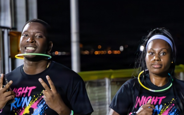 Two runners participating in a nighttime fun run event organized by MWR Fitness, wearing matching 'Night Run' themed black t-shirts with colorful paint splatter designs. Runner on the left wears bib number 75 and is holding a smartphone while making a peace sign, with a glow stick in his mouth. Runner on the right wears bib number 76 and has glow stick accessories around her neck and wrist.
