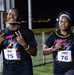 Two runners participating in a nighttime fun run event organized by MWR Fitness, wearing matching 'Night Run' themed black t-shirts with colorful paint splatter designs. Runner on the left wears bib number 75 and is holding a smartphone while making a peace sign, with a glow stick in his mouth. Runner on the right wears bib number 76 and has glow stick accessories around her neck and wrist.