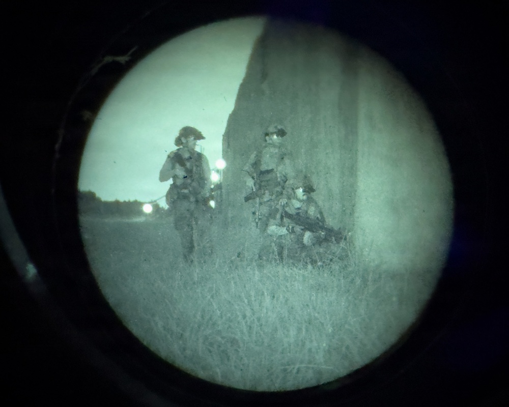 Image captured through a night vision optic showing three armed military or tactical personnel positioned near a large structure or wall at dusk/night. The circular vignette and green-gray tonal rendering are characteristic of night vision device (NVD) imagery. Personnel appear to be conducting a tactical movement or security operation in an open grassy area.