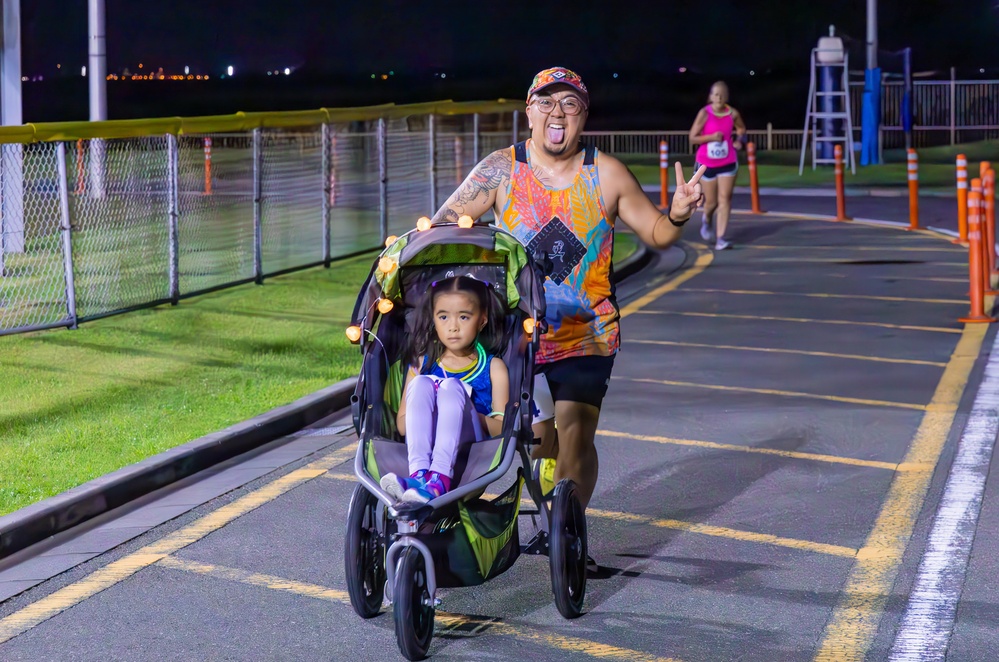 A nighttime running race event where a man in a colorful tank top pushes a young girl in a jogging stroller while making a peace sign and sticking out his tongue. The scene takes place on a paved track or path lined with orange safety cones and a chain-link fence. A female runner in a pink top is visible in the background also participating in the race.