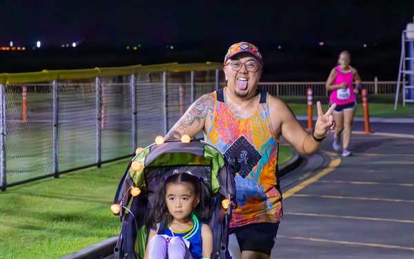 A nighttime running race event where a man in a colorful tank top pushes a young girl in a jogging stroller while making a peace sign and sticking out his tongue. The scene takes place on a paved track or path lined with orange safety cones and a chain-link fence. A female runner in a pink top is visible in the background also participating in the race.
