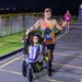 A nighttime running race event where a man in a colorful tank top pushes a young girl in a jogging stroller while making a peace sign and sticking out his tongue. The scene takes place on a paved track or path lined with orange safety cones and a chain-link fence. A female runner in a pink top is visible in the background also participating in the race.