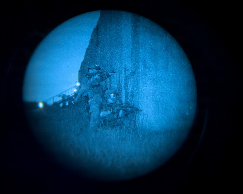 The image shows what appears to be an underwater shipwreck photographed through a circular porthole or submersible viewport. The wreck is heavily deteriorated and encrusted, resting at a steep angle on the seafloor. Artificial lighting from what appears to be a submersible or ROV illuminates the scene in a deep blue hue.