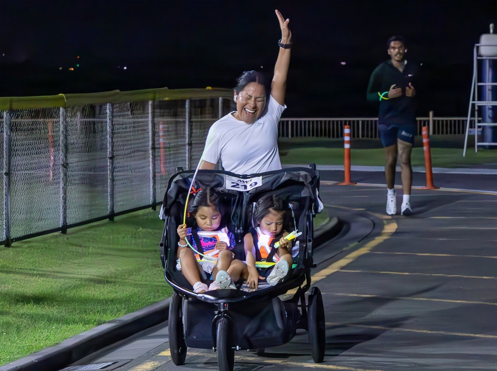 A woman wearing bib number 219 celebrates with one arm raised while pushing a double jogging stroller carrying two young children during a nighttime race event. The children are holding glow sticks and wearing safety vests, while the runner appears joyful, likely crossing or approaching the finish line. A second runner wearing a Nike outfit is visible in the background.
