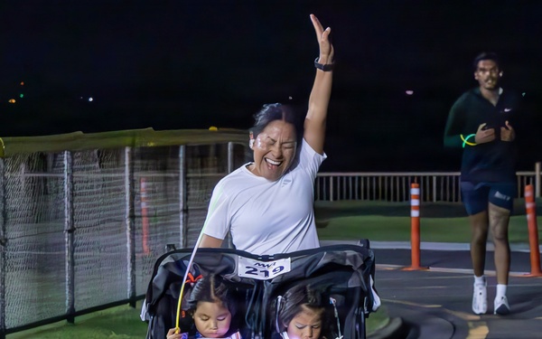 A woman wearing bib number 219 celebrates with one arm raised while pushing a double jogging stroller carrying two young children during a nighttime race event. The children are holding glow sticks and wearing safety vests, while the runner appears joyful, likely crossing or approaching the finish line. A second runner wearing a Nike outfit is visible in the background.