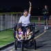 A woman wearing bib number 219 celebrates with one arm raised while pushing a double jogging stroller carrying two young children during a nighttime race event. The children are holding glow sticks and wearing safety vests, while the runner appears joyful, likely crossing or approaching the finish line. A second runner wearing a Nike outfit is visible in the background.