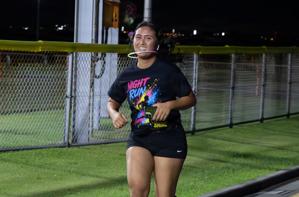 A young woman is running at night during what appears to be an organized 'Night Run' event. She is wearing a black graphic t-shirt that reads 'Night Run' and is accessorized with a glow ring around her neck. The photo is taken outdoors near a chain-link fence with artificial turf visible in the background.