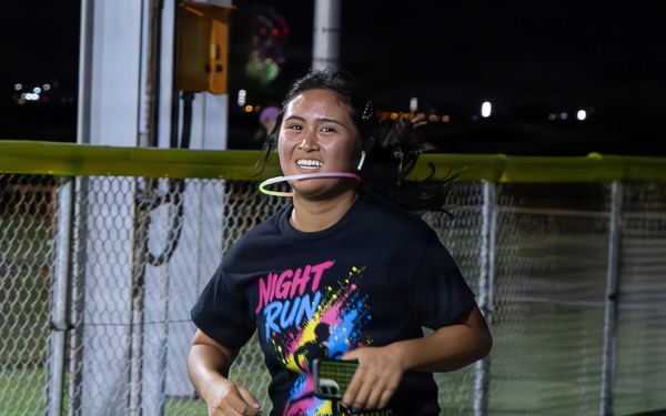 A young woman is running at night during what appears to be an organized 'Night Run' event. She is wearing a black graphic t-shirt that reads 'Night Run' and is accessorized with a glow ring around her neck. The photo is taken outdoors near a chain-link fence with artificial turf visible in the background.