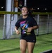A young woman is running at night during what appears to be an organized 'Night Run' event. She is wearing a black graphic t-shirt that reads 'Night Run' and is accessorized with a glow ring around her neck. The photo is taken outdoors near a chain-link fence with artificial turf visible in the background.