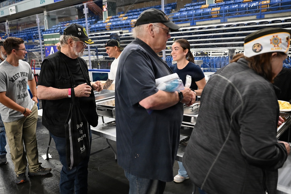 A group of people are gathered at what appears to be a food service or community meal event inside a large arena or stadium venue. Volunteers and attendees are interacting along a buffet-style serving line with chafing dishes. Several individuals are wearing military or veterans organization caps, suggesting this may be a veterans-related event.