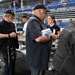 A group of people are gathered at what appears to be a food service or community meal event inside a large arena or stadium venue. Volunteers and attendees are interacting along a buffet-style serving line with chafing dishes. Several individuals are wearing military or veterans organization caps, suggesting this may be a veterans-related event.