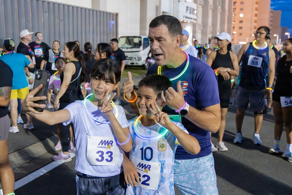 A group of participants at an MWR (Morale, Welfare, and Recreation) fitness run event, likely a glow run given the neon glow sticks and bracelets visible. Two children wearing race bibs numbered 33 and 62 are taking a selfie with an adult man, all making hand gestures and smiling. The event appears to be taking place at dusk or nighttime in an outdoor urban or military base setting.