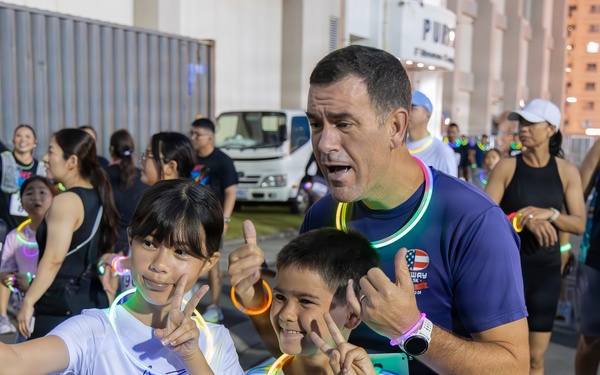 A group of participants at an MWR (Morale, Welfare, and Recreation) fitness run event, likely a glow run given the neon glow sticks and bracelets visible. Two children wearing race bibs numbered 33 and 62 are taking a selfie with an adult man, all making hand gestures and smiling. The event appears to be taking place at dusk or nighttime in an outdoor urban or military base setting.