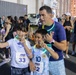 A group of participants at an MWR (Morale, Welfare, and Recreation) fitness run event, likely a glow run given the neon glow sticks and bracelets visible. Two children wearing race bibs numbered 33 and 62 are taking a selfie with an adult man, all making hand gestures and smiling. The event appears to be taking place at dusk or nighttime in an outdoor urban or military base setting.