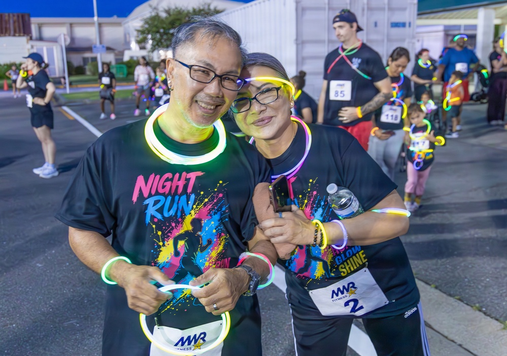 Two smiling participants pose together at a Night Run glow race event, wearing matching black 'Night Run' t-shirts and decorated with colorful glow sticks and necklaces. They are wearing MWR Fitness race bibs (numbers visible) and appear to be at the starting area of the event in a parking lot during evening hours. Multiple other participants in similar attire are visible in the background.