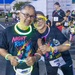 Two smiling participants pose together at a Night Run glow race event, wearing matching black 'Night Run' t-shirts and decorated with colorful glow sticks and necklaces. They are wearing MWR Fitness race bibs (numbers visible) and appear to be at the starting area of the event in a parking lot during evening hours. Multiple other participants in similar attire are visible in the background.