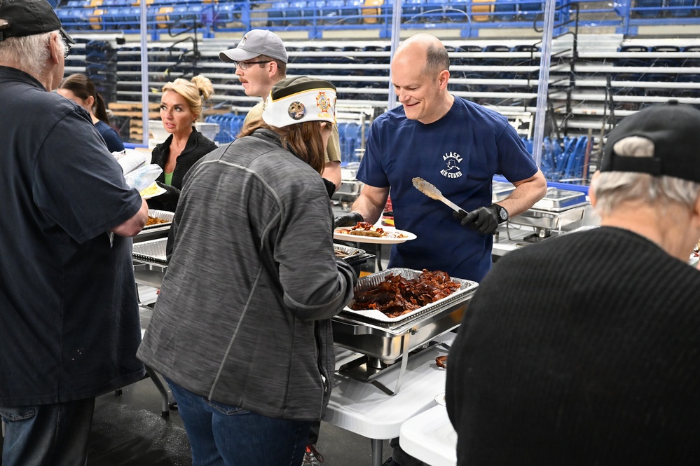 A food service event is taking place inside what appears to be a large indoor arena or sports facility. A man wearing an 'Alaska Air Guard' t-shirt and black gloves is serving food from chafing dishes to attendees. The event appears to be a community or military appreciation meal, with several volunteers and guests visible.