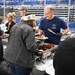A food service event is taking place inside what appears to be a large indoor arena or sports facility. A man wearing an 'Alaska Air Guard' t-shirt and black gloves is serving food from chafing dishes to attendees. The event appears to be a community or military appreciation meal, with several volunteers and guests visible.