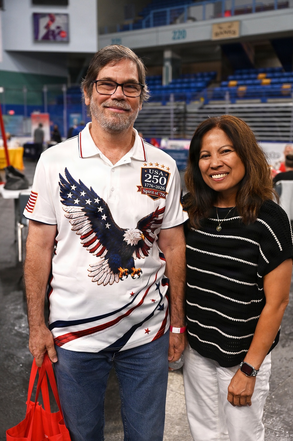 Two adults pose together smiling inside what appears to be an indoor arena or sports venue. The man on the left wears a patriotic USA 250th anniversary shirt (1776-2026) featuring a bald eagle design, while the woman on the right wears a black and white striped knit top. The background shows blue stadium seating and arena infrastructure.