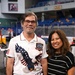 Two adults pose together smiling inside what appears to be an indoor arena or sports venue. The man on the left wears a patriotic USA 250th anniversary shirt (1776-2026) featuring a bald eagle design, while the woman on the right wears a black and white striped knit top. The background shows blue stadium seating and arena infrastructure.