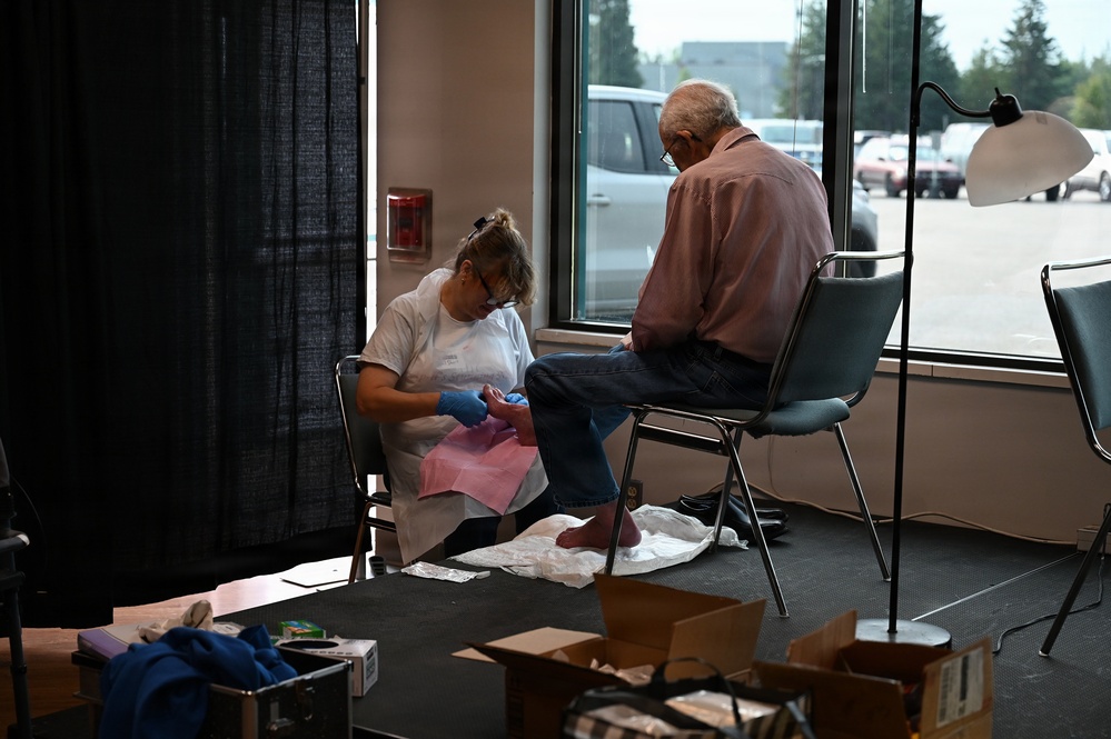 A healthcare worker wearing blue gloves and a white uniform is providing foot care to an elderly man seated in a chair near a large window. The setting appears to be a temporary or community health clinic set up in a large open space, possibly a community center or converted retail space. Medical supplies and boxes are visible on the floor nearby.