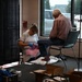 A healthcare worker wearing blue gloves and a white uniform is providing foot care to an elderly man seated in a chair near a large window. The setting appears to be a temporary or community health clinic set up in a large open space, possibly a community center or converted retail space. Medical supplies and boxes are visible on the floor nearby.