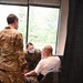 Two military personnel in OCP (Operational Camouflage Pattern) uniforms are attending to a civilian man seated in a folding chair. Both service members are wearing blue face masks, suggesting a COVID-19 era medical or assistance context. The interaction appears to involve a physical assessment or medical assistance of the seated elderly man.