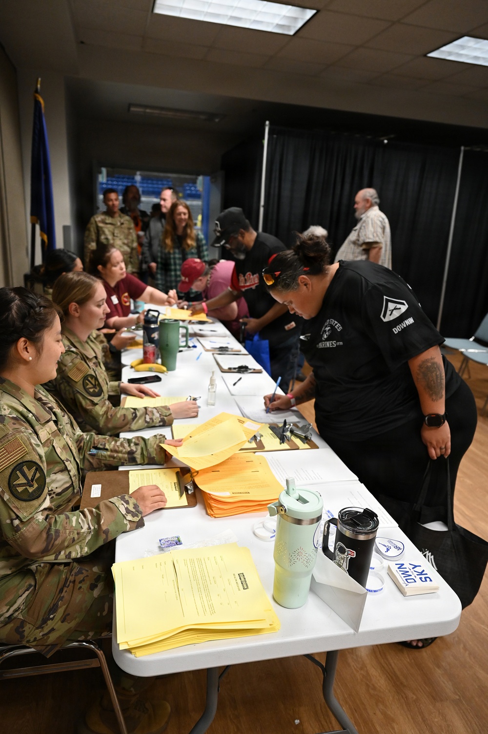 Military personnel in camouflage uniforms are seated at a long white table processing paperwork for civilians and veterans at what appears to be a registration or intake event. Multiple people are filling out forms on clipboards, with stacks of yellow and orange documents visible on the table. The setting appears to be an indoor facility, possibly a community center or military installation.