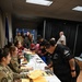 Military personnel in camouflage uniforms are seated at a long white table processing paperwork for civilians and veterans at what appears to be a registration or intake event. Multiple people are filling out forms on clipboards, with stacks of yellow and orange documents visible on the table. The setting appears to be an indoor facility, possibly a community center or military installation.