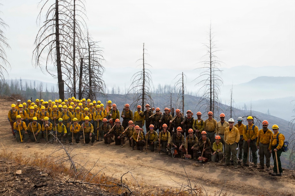 A large group of wildland firefighters poses for a group photo on a hillside surrounded by burned, charred trees. The crew is dressed in standard wildland firefighting gear including yellow Nomex shirts and hard hats in yellow and orange. The smoky, hazy background reveals a fire-affected landscape with scorched terrain and distant smoke-filled valleys.