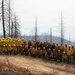 A large group of wildland firefighters poses for a group photo on a hillside surrounded by burned, charred trees. The crew is dressed in standard wildland firefighting gear including yellow Nomex shirts and hard hats in yellow and orange. The smoky, hazy background reveals a fire-affected landscape with scorched terrain and distant smoke-filled valleys.