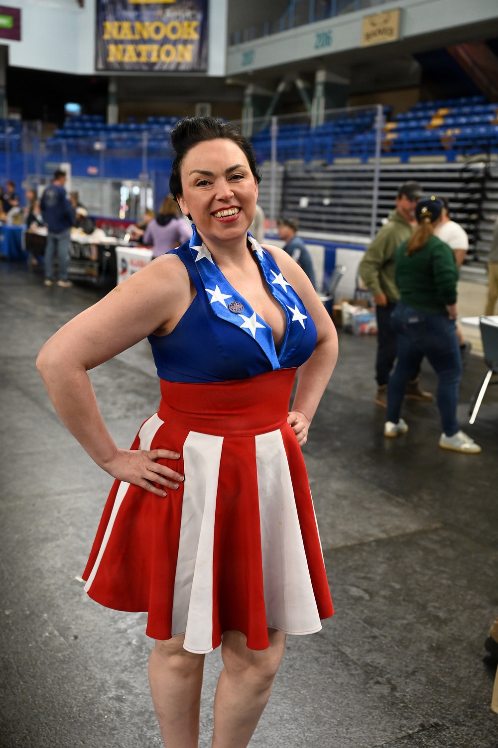A woman poses confidently inside what appears to be the Carlson Center arena at the University of Alaska Fairbanks, home of the Nanooks. She is wearing a patriotic American flag-themed dress with a blue bodice featuring white stars and a red-and-white striped skirt, along with a small 'I Voted' style button. The setting appears to be an indoor event or convention taking place on the arena floor.
