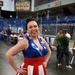 A woman poses confidently inside what appears to be the Carlson Center arena at the University of Alaska Fairbanks, home of the Nanooks. She is wearing a patriotic American flag-themed dress with a blue bodice featuring white stars and a red-and-white striped skirt, along with a small 'I Voted' style button. The setting appears to be an indoor event or convention taking place on the arena floor.