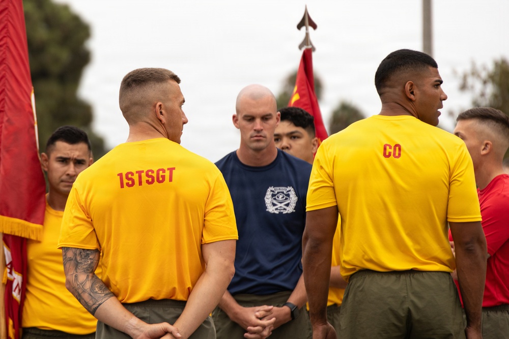 A group of military personnel, likely U.S. Marines, are gathered outdoors in what appears to be a formation or briefing. Two individuals in yellow t-shirts with 'CO' and '1STSGT' printed on the back are prominently visible, indicating company commander and first sergeant roles. A Marine in a navy blue t-shirt with a unit insignia stands facing them, possibly addressing the group.