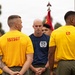A group of military personnel, likely U.S. Marines, are gathered outdoors in what appears to be a formation or briefing. Two individuals in yellow t-shirts with 'CO' and '1STSGT' printed on the back are prominently visible, indicating company commander and first sergeant roles. A Marine in a navy blue t-shirt with a unit insignia stands facing them, possibly addressing the group.